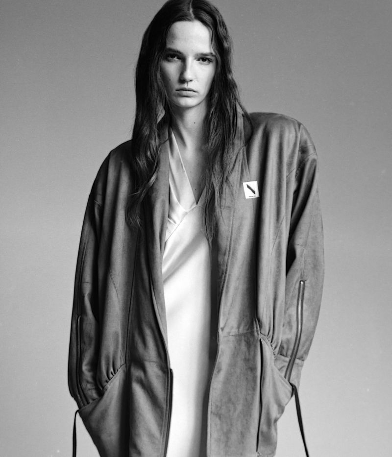 Model Emma Muszela wearing a long oversized jacket and light slip dress against a grey studio backdrop by Thomas Daloiso.