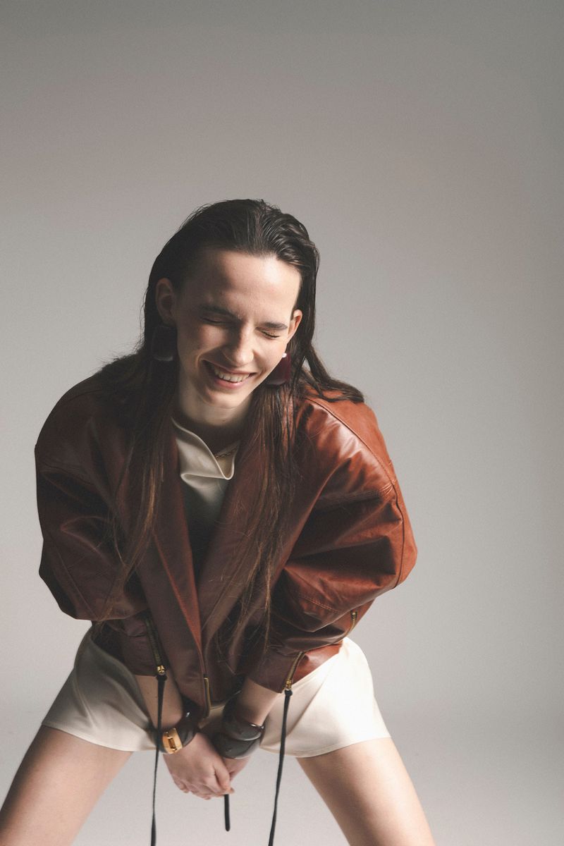 Smiling female model in a brown leather jacket and cream shorts posing against a neutral grey studio seamless backdrop.
