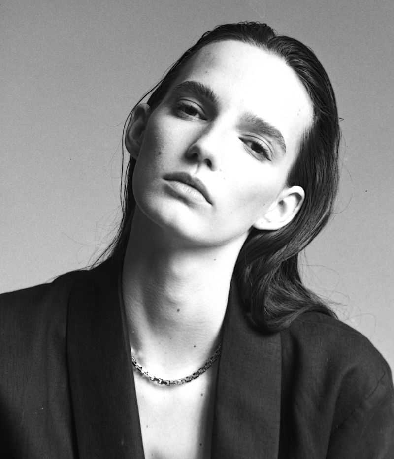 Black and white portrait of a model wearing a dark blazer and a link chain necklace against a light grey background.