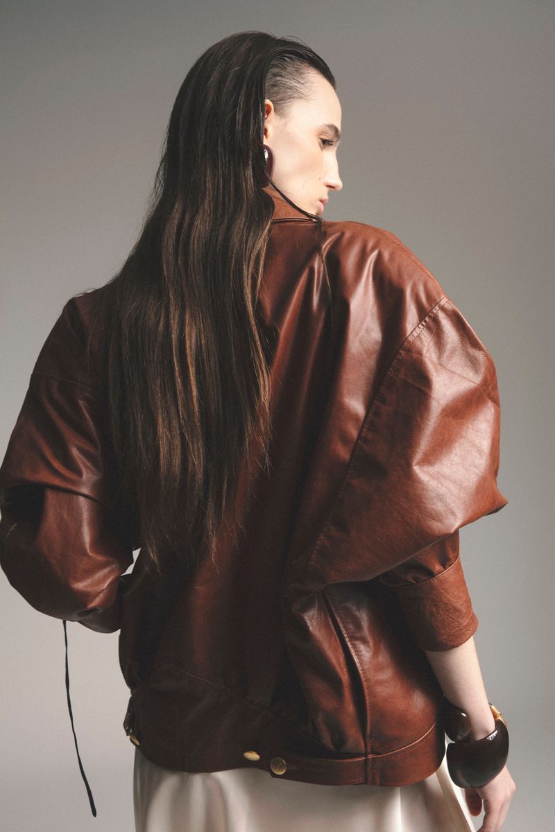 Model with long dark hair wearing a brown leather jacket and light skirt against a grey background