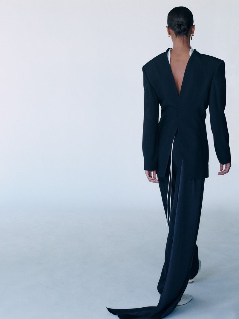 Back view of an adult female walking away, wearing a black blazer with tail detail, in a bright, neutral studio with minimal styling.