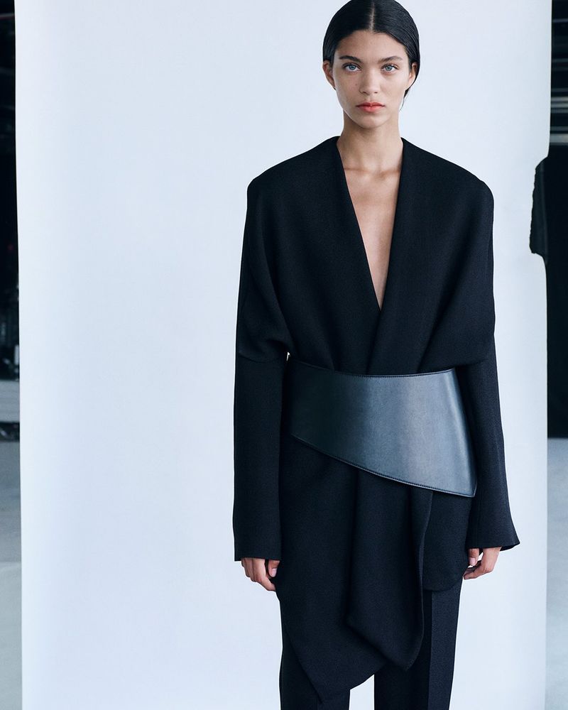 Model wearing a black belted coat and trousers against a white seamless background for Tove