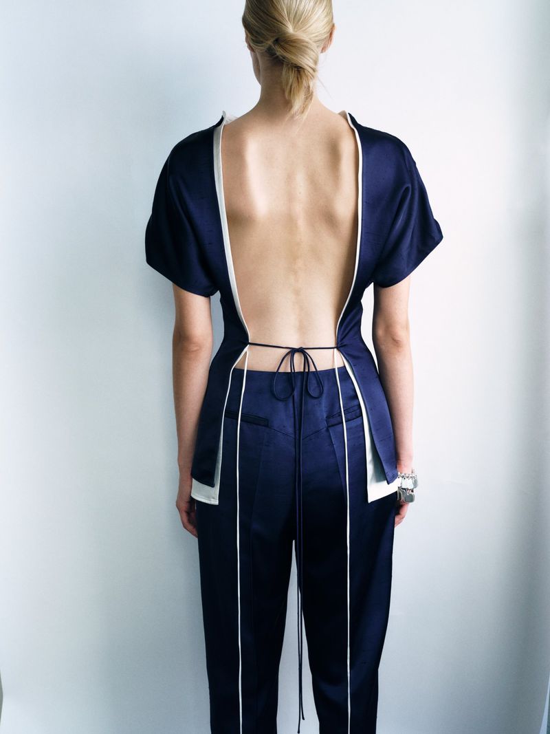 Model seen from behind wearing a navy blue open-back top with white piping and matching trousers against a white seamless backdrop.