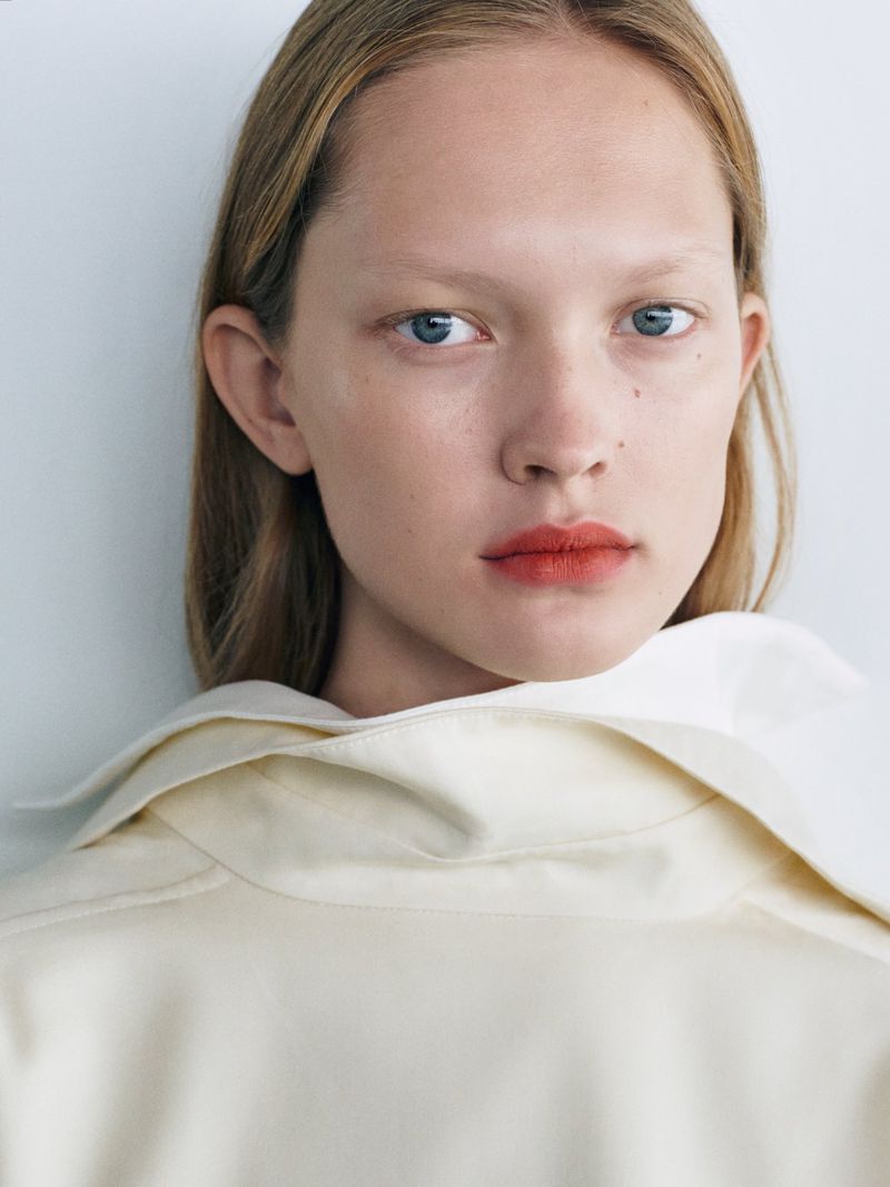 Close-up portrait of a model with blonde hair wearing a cream-colored high-collared garment against a light grey background.