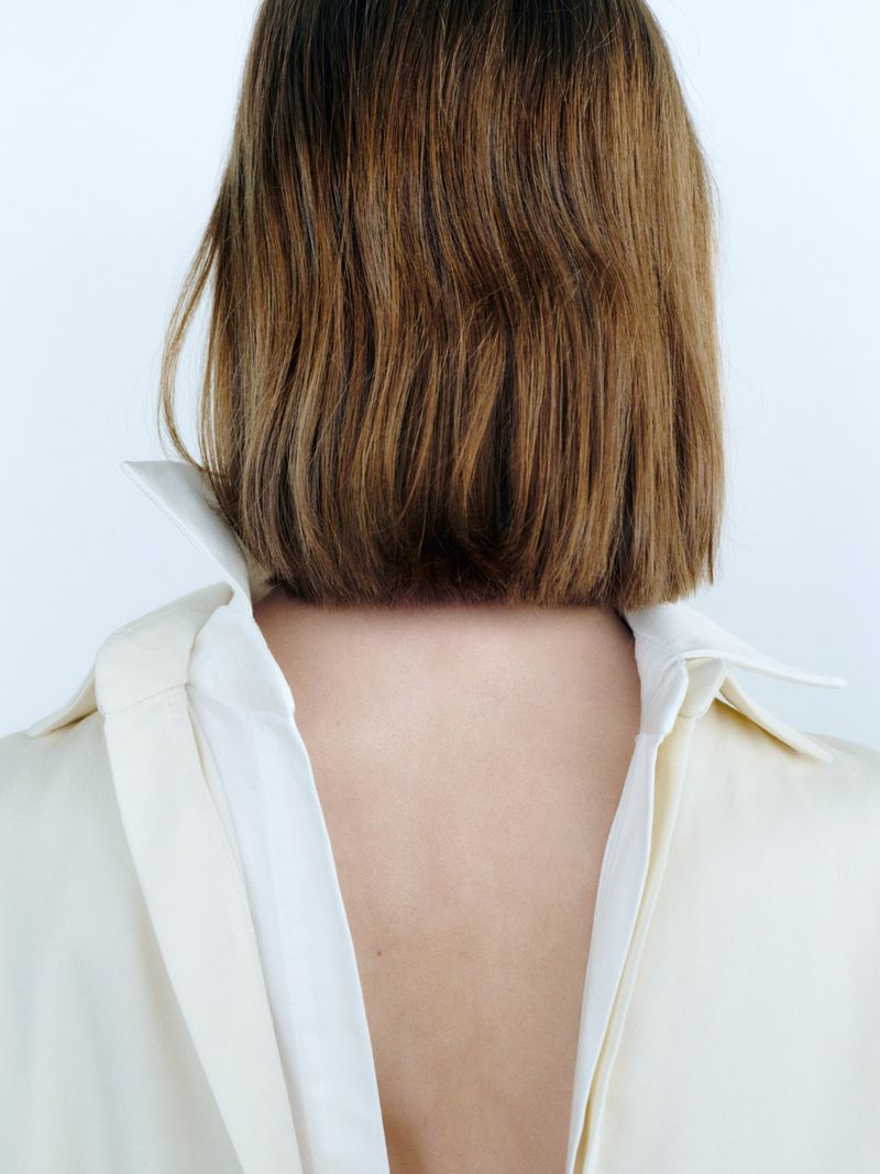 Close-up back view of a person with shoulder-length brown bob hair wearing a cream-colored collared shirt against a white wall.