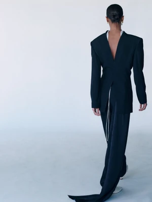 Model seen from behind wearing a black structured jacket and trousers against a pale studio background.