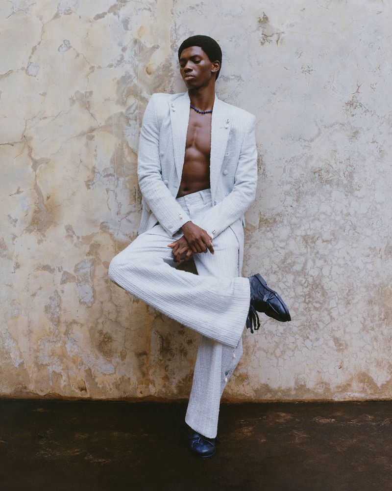 Model wearing a light grey suit jacket and trousers posing against a weathered textured wall for Byredo Bal d’Afrique 2026.