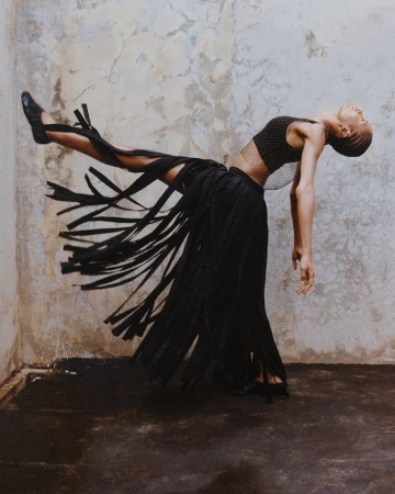 A slender female model in a black fringed skirt and top strikes a dynamic pose indoors against a mottled wall, exuding a sense of movement.