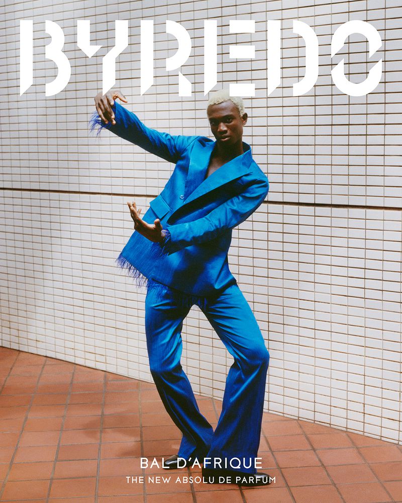 Black man with dyed blonde hair wearing a bright blue suit posing dynamically against a white tiled wall