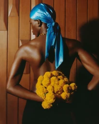 Model wearing bright blue satin durag holding yellow flowers against wooden paneling