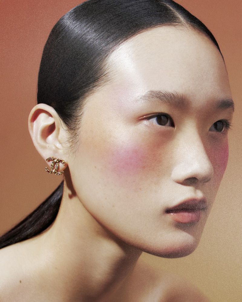 Close-up portrait of an Asian woman with slicked-back hair and a gold Chanel earring, against a softly textured peach background.