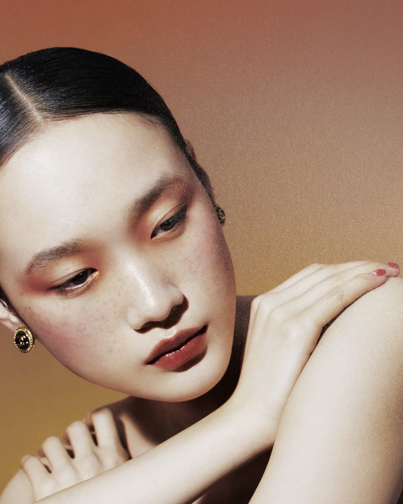 Close-up beauty shot of an Asian female model with clean makeup and styled hair, wearing gold earrings, against a gradient background.