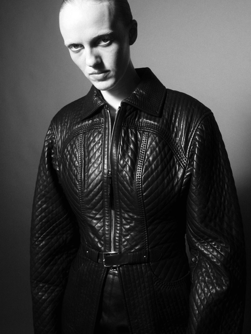 Model wearing a black quilted leather jacket with a belt against a grey studio backdrop in a black and white portrait.