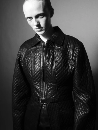 Model wearing a black quilted leather jacket with a belt against a grey studio backdrop in a black and white portrait.