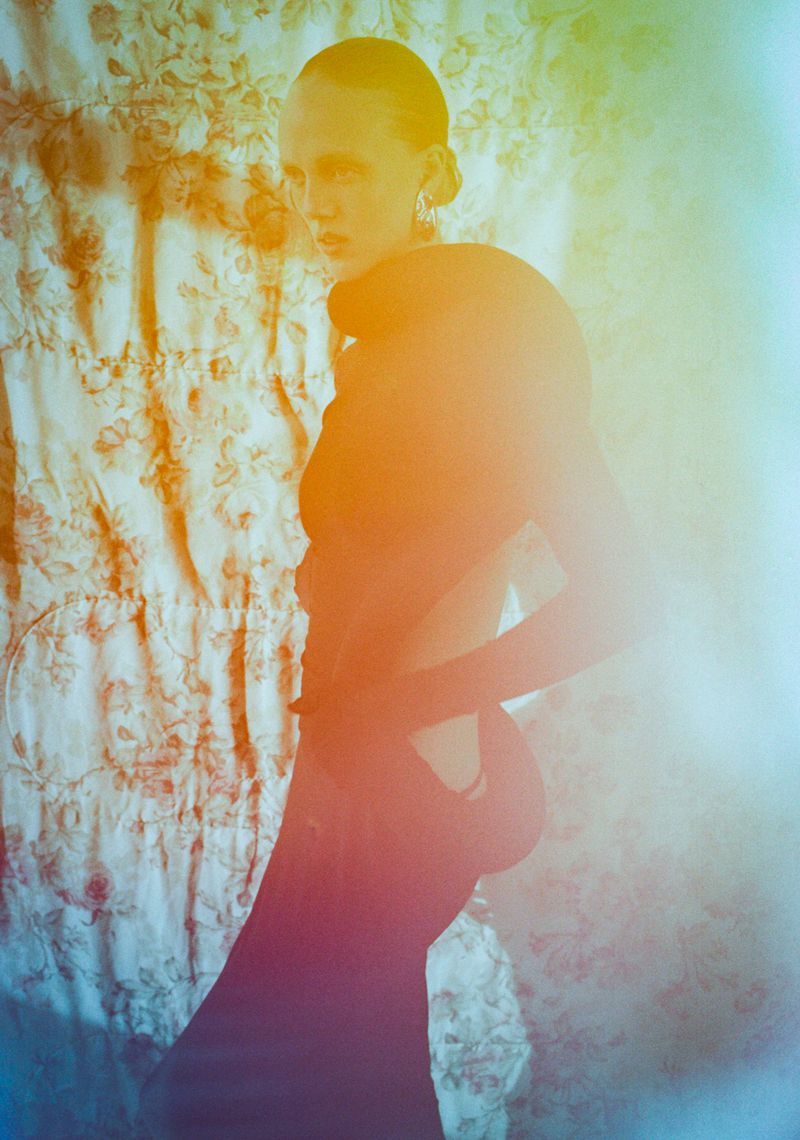 Model in a dark sculptural top standing against a patterned floral curtain with strong color grading
