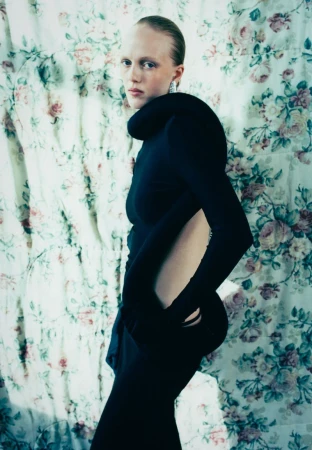 Model in black sculptural dress standing in profile against a floral patterned fabric background for Cero Magazine BOY project.