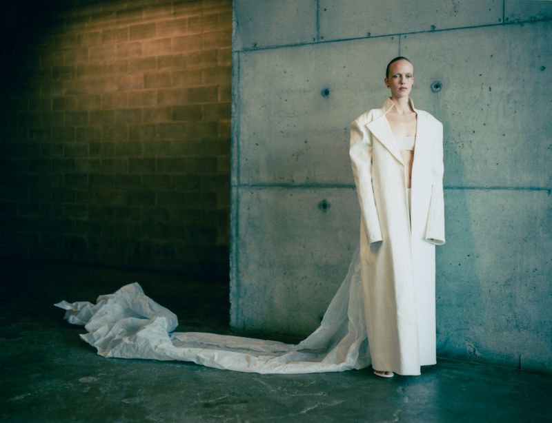 An adult woman in a white coat and a bra top poses against a concrete wall indoors with a soft light and a long white train.