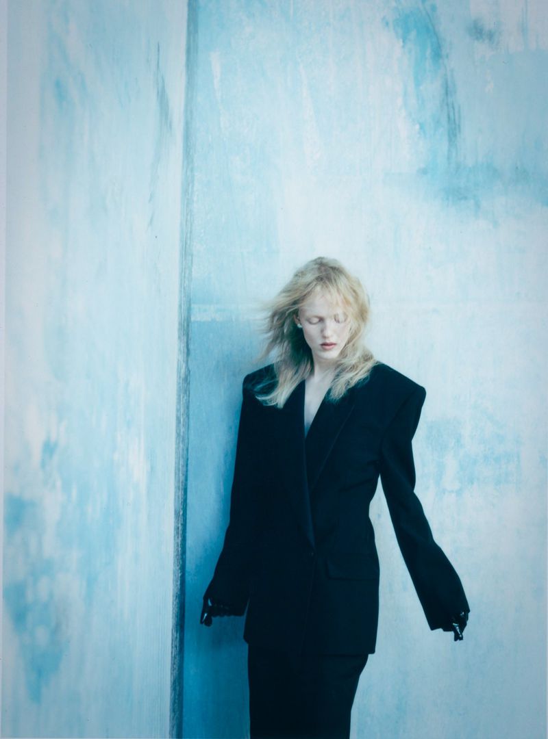 Model with blonde hair wearing a structured black blazer against a pale blue textured wall in an editorial fashion photograph.