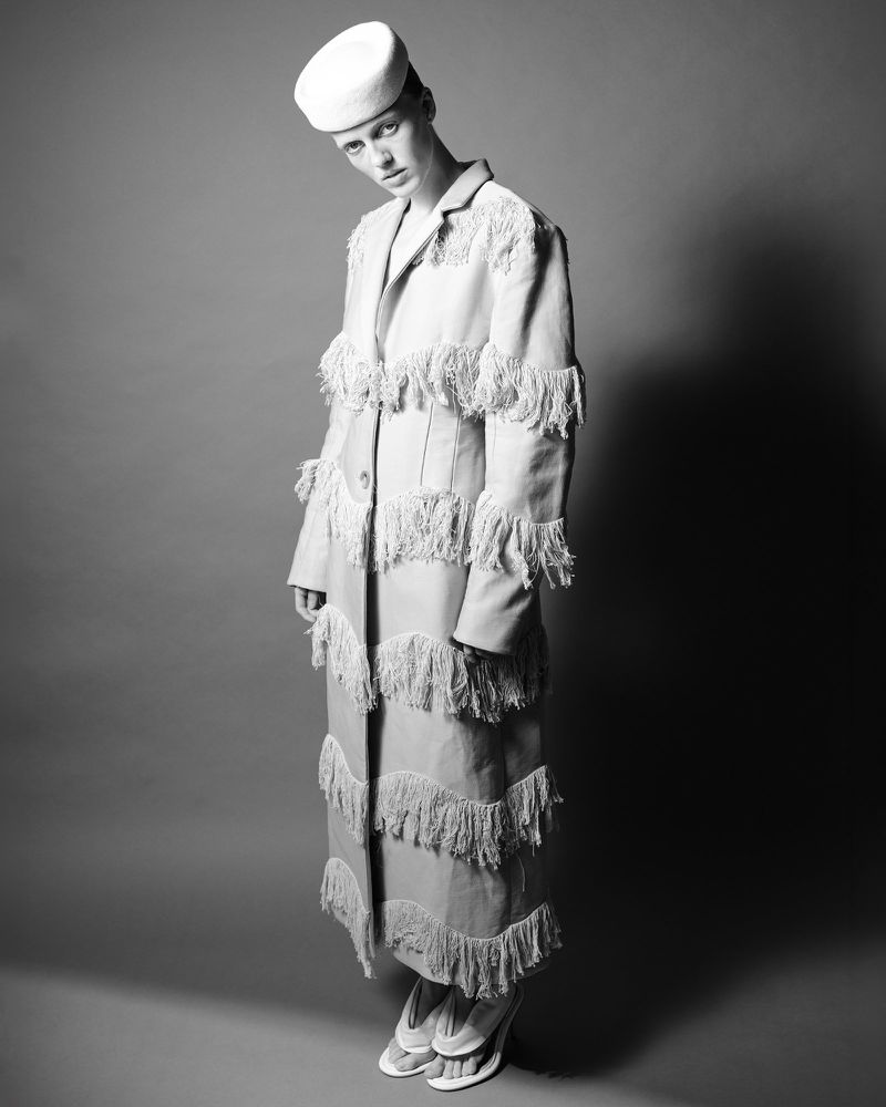 Model wearing a fringed coat and pillbox hat standing against a grey seamless background