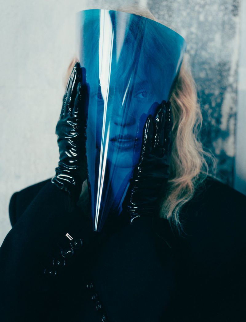Portrait of a person holding a blue conical visor and wearing black latex gloves and a dark jacket against a textured wall.