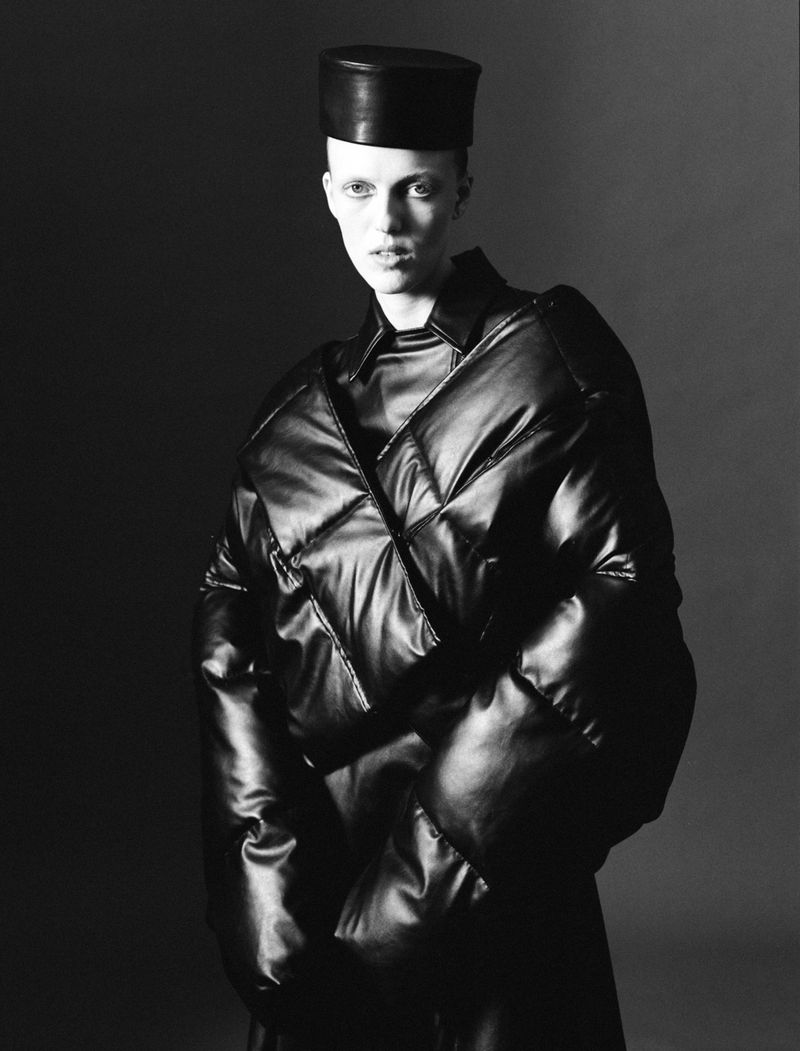 Model wearing a structured black leather puffer jacket and pillbox hat against a dark studio background