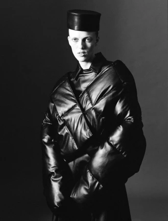 Model wearing a structured black leather puffer jacket and pillbox hat against a dark studio background