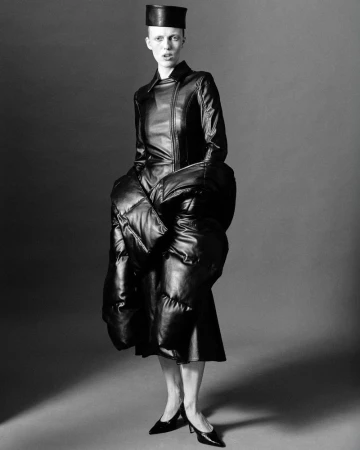 Model wearing black leather coat and structured pillbox hat standing against a grey studio backdrop for Cero Magazine.