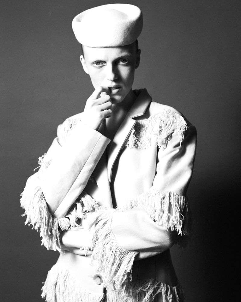 Model wearing a fringed jacket and pillbox hat with finger near mouth in a black and white portrait