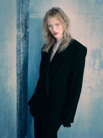 Model with blonde hair wearing an oversized black blazer and matching trousers against a textured blue concrete wall.