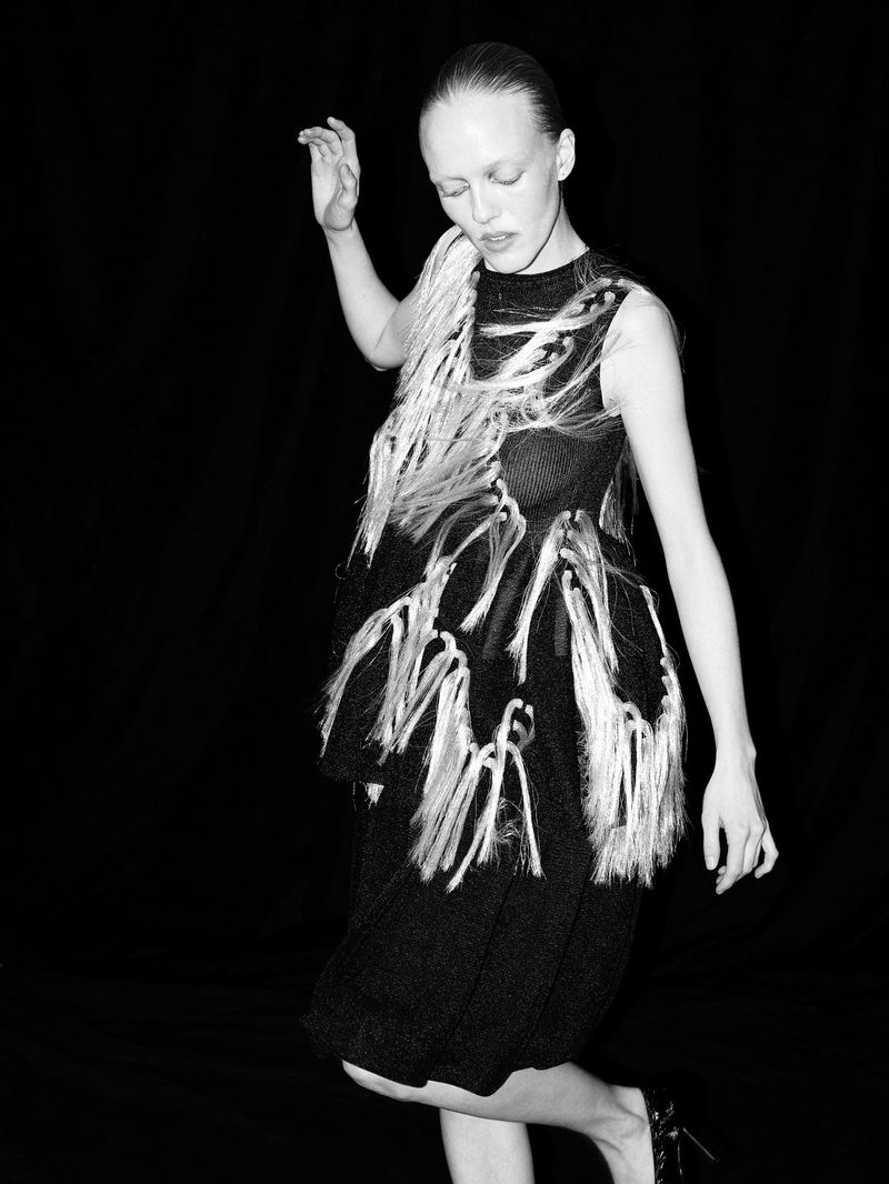 Model in a black textured dress with light fringe details against a black background