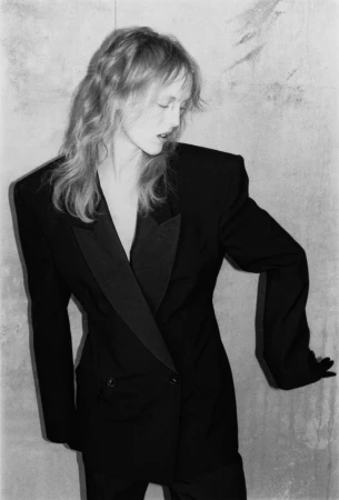 Model wearing a black tailored blazer with padded shoulders against a textured grey wall