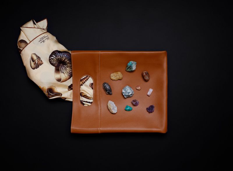 Tan leather pouch with cutout handles and a cream silk scarf featuring seashell prints, displayed with small crystals on a black background.