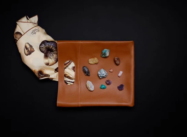 Tan leather pouch with cutout handles and a cream silk scarf featuring seashell prints, displayed with small crystals on a black background.