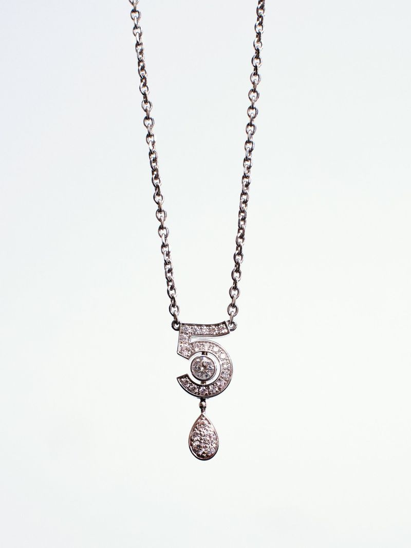 Close-up product shot of a silver diamond-pave number five pendant necklace hanging against a pale background.