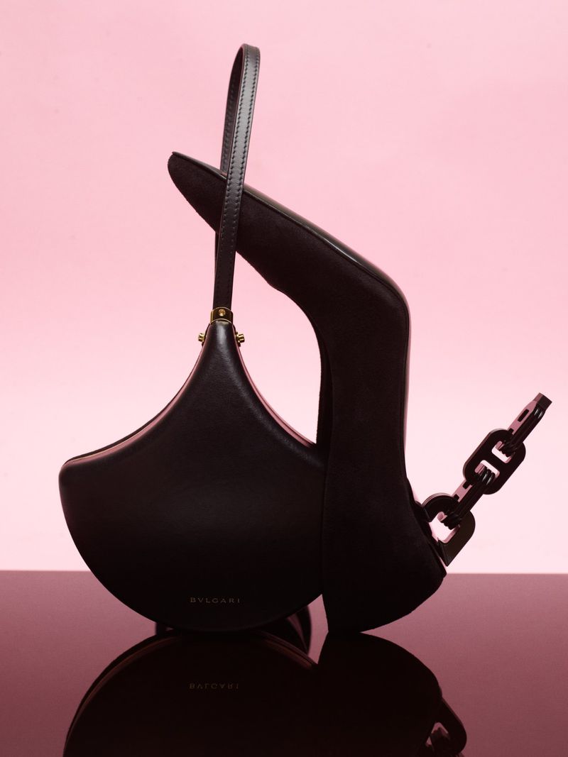 Black sculptural leather handbag with handle and chain detail positioned on a reflective surface against a pink backdrop