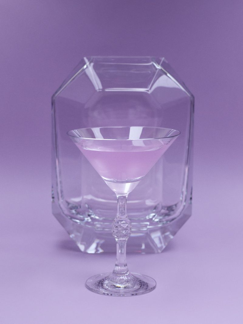 A crystal martini glass with pink liquid in front of a faceted glass block on a solid purple background