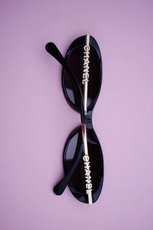 Black sunglasses with metallic branding on temples displayed vertically against a pink background