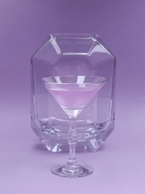 A crystal martini glass with pink liquid in front of a faceted glass block on a solid purple background
