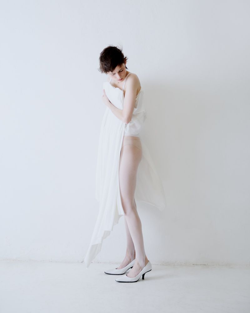 Model wrapped in white fabric wearing white heels posing against a plain white background for The Greatest Magazine.