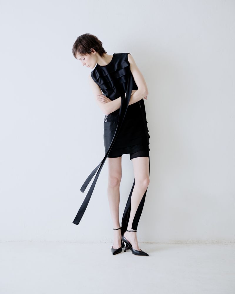 Model in black dress with long hanging straps posing against a plain white studio background.