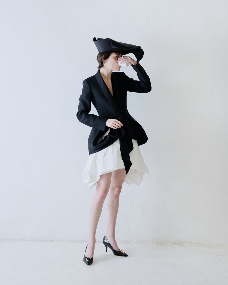 Model in black jacket and white skirt holding small cup against a white backdrop