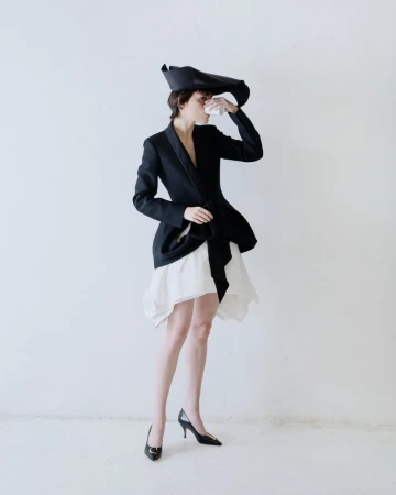 Model in black jacket and white skirt holding small cup against a white backdrop