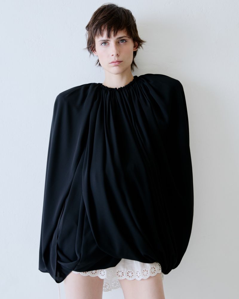 Model in a voluminous black top and white lace shorts standing against a plain white wall