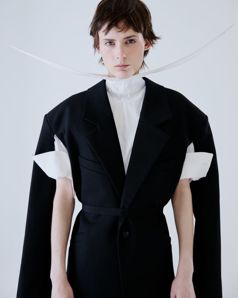 Model wearing a black blazer over a white shirt with a white ribbon against a plain light grey background for The Greatest Magazine.