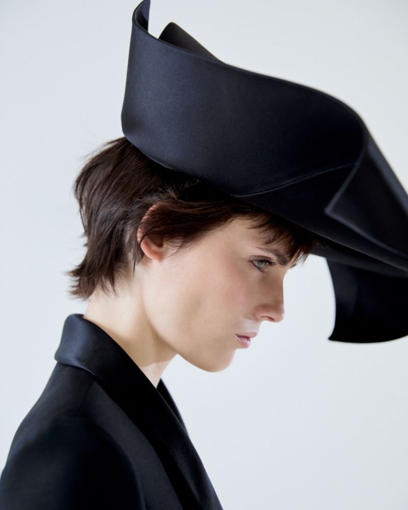 Profile portrait of a woman wearing an oversized black sculptural hat and black blazer against a white seamless background.