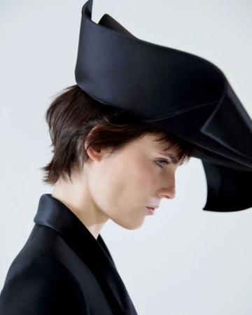 Profile portrait of a woman wearing an oversized black sculptural hat and black blazer against a white seamless background.