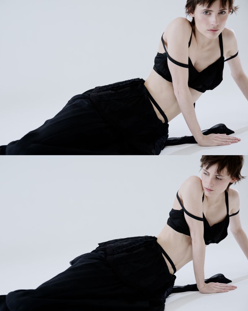Woman in a black two-piece outfit poses in a studio. The outfit features a bra top and loose trousers. She's kneeling on a white surface.
