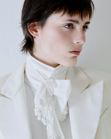 Model with dark short hair wearing a white blazer and high-necked laced scarf against a neutral background.