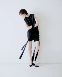 Model in black dress with long hanging straps posing against a plain white studio background.