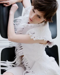 Model with short brown hair wearing a white textured fringed garment seated against a black chair and white background